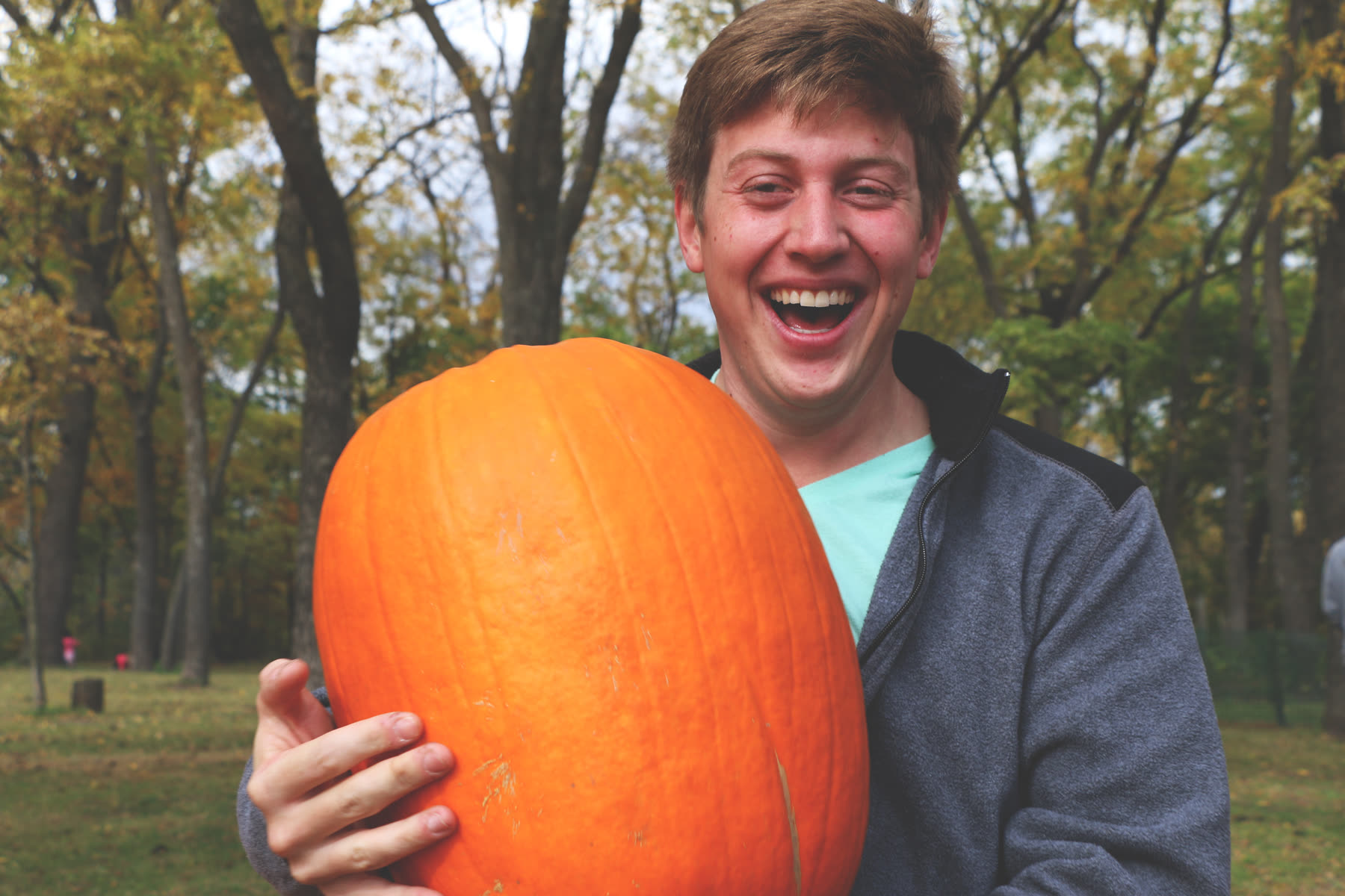 A visitor laughing with a big pumpkin under the trees